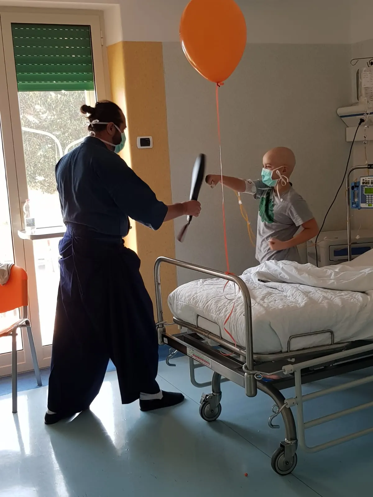 Un copil cu masca loveste o paleta de antrenament langa patul de spital, in timp ce un balon pluteste deasupra.