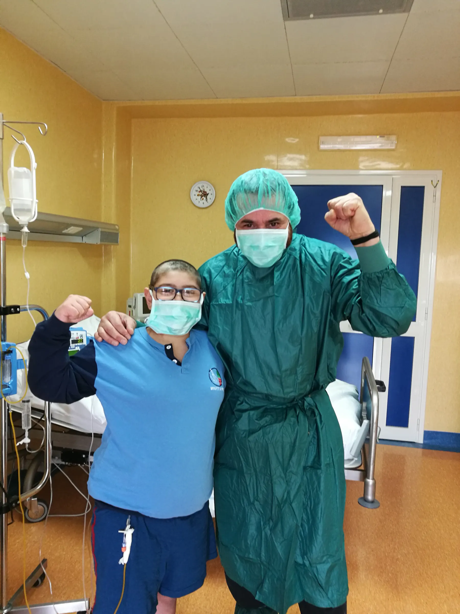 Un copil cu masca si un instructor in echipament de protectie ridica pumnii intr-un salon de spital.