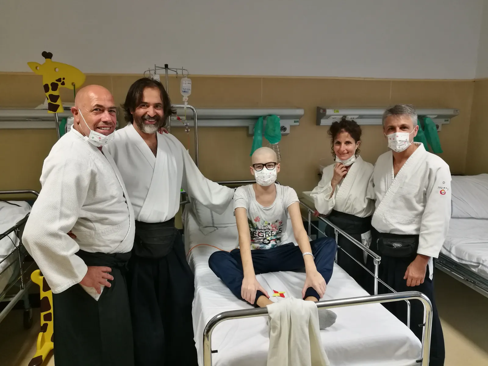 Mai multi voluntari in echipament Budo stau in jurul unui pacient cu masca, asezat pe patul de spital.