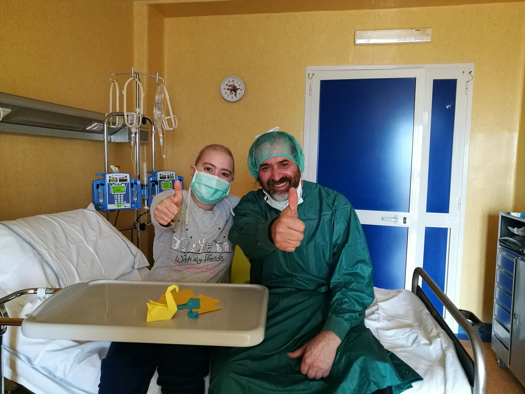 Un copil cu masca si un instructor in echipament medical arata semnul de aprobare intr-un salon de spital.